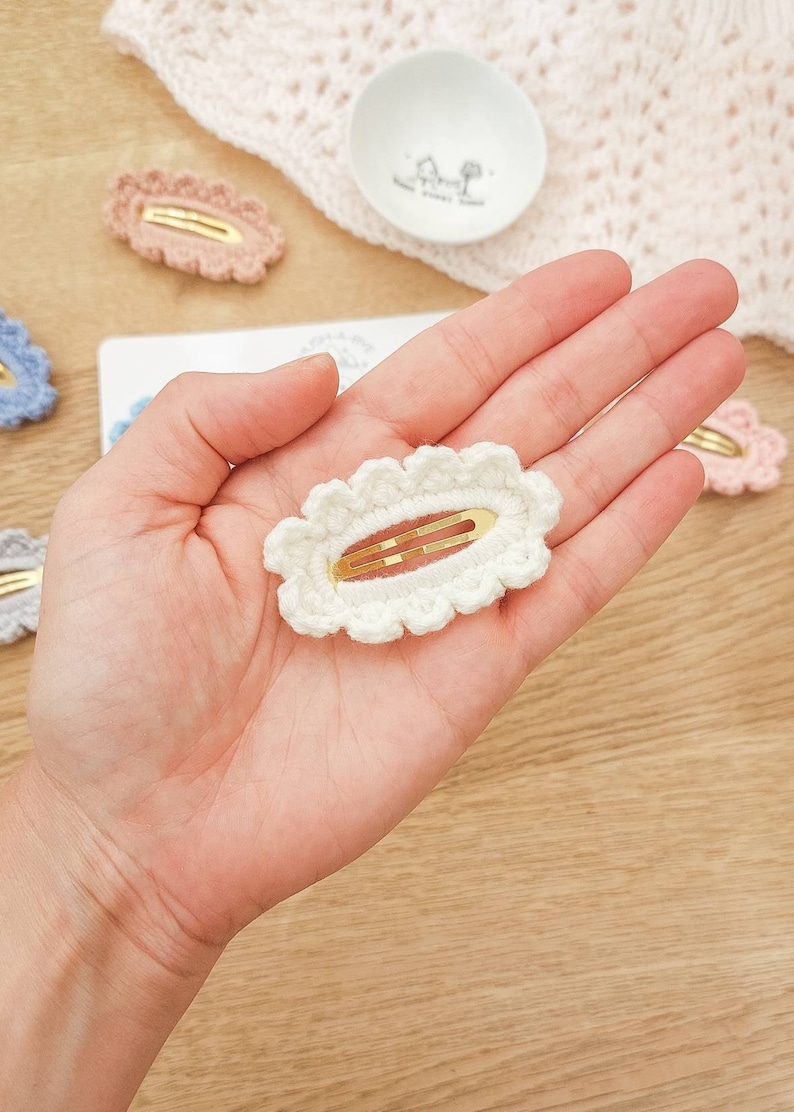 May include: A white crocheted hair clip with a gold metal bar. The clip is oval shaped with a scalloped edge.