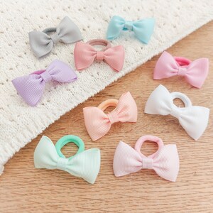 May include: A collection of eight hair ties featuring small, ribbed fabric bows in various pastel colours including light blue, grey, lavender, pink, peach, white, mint green, and soft blush, each attached to a matching elastic hair band.