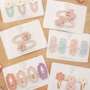 May include: A set of handmade crocheted hair clips in various colors and styles, including flowers, bows, and oval shapes. The clips are displayed on white cards with the text "hushabye bay" and a small illustration of a bear.