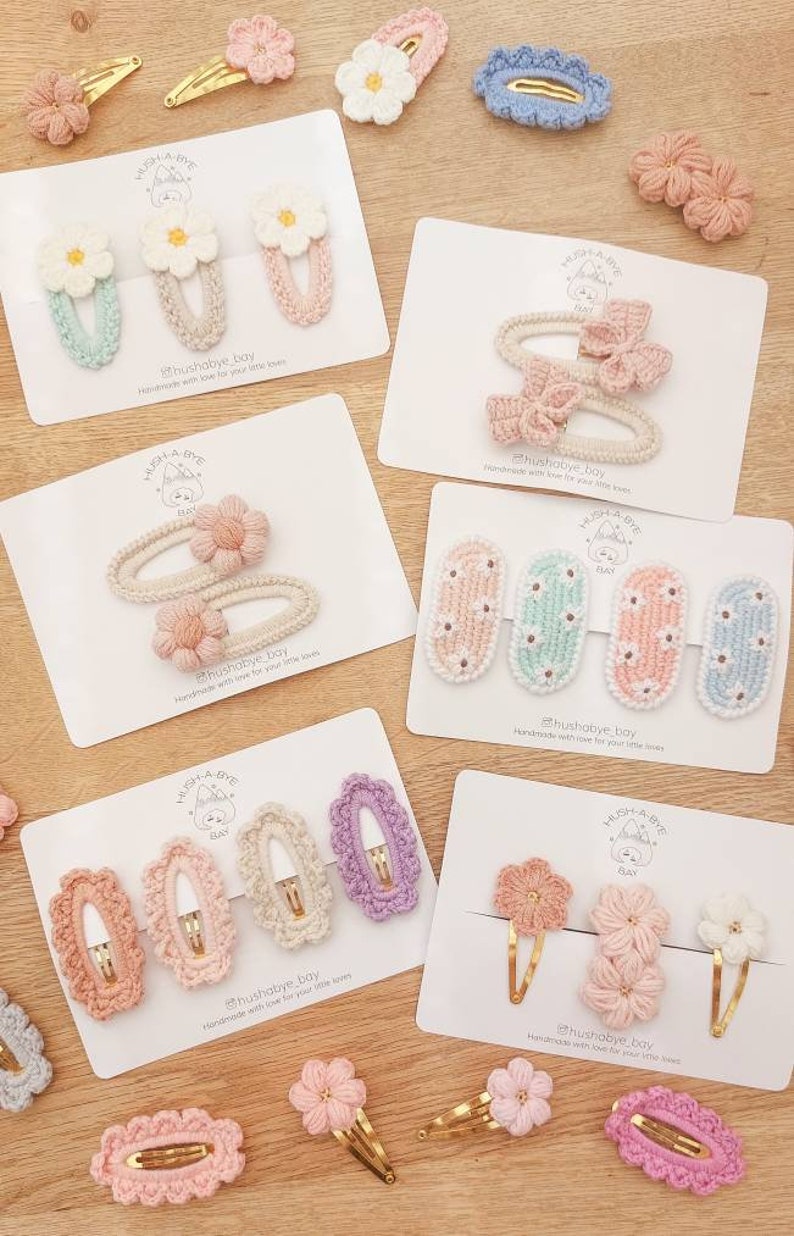May include: A set of handmade crocheted hair clips in various colors and styles, including flowers, bows, and oval shapes. The clips are displayed on white cards with the text "hushabye bay" and a small illustration of a bear.
