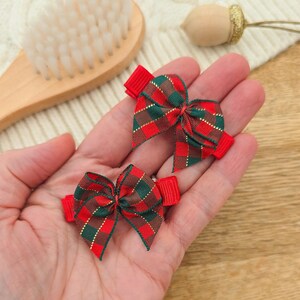 Puede incluir: Dos peque&ntilde;os lazos a cuadros rojos y verdes con clips de cinta roja. Los lazos se sostienen en la mano de una persona. Los lazos tienen un hilo met&aacute;lico dorado tejido en el patr&oacute;n de cuadros. Un cepillo de madera y una bellota decorativa est&aacute;n en el fondo.