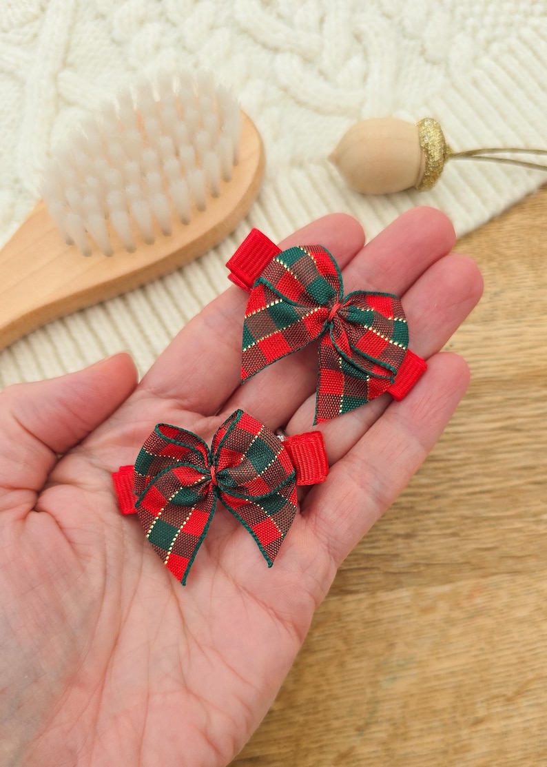 Puede incluir: Dos peque&ntilde;os lazos a cuadros rojos y verdes con clips de cinta roja. Los lazos se sostienen en la mano de una persona. Los lazos tienen un hilo met&aacute;lico dorado tejido en el patr&oacute;n de cuadros. Un cepillo de madera y una bellota decorativa est&aacute;n en el fondo.