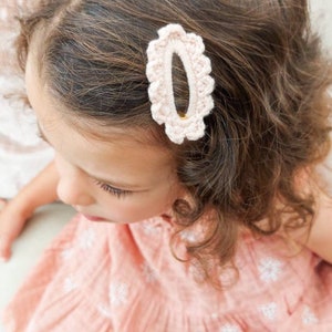 May include: A young girl with curly brown hair wearing a pink dress with white floral embroidery. She has a pink crocheted hair clip in her hair.
