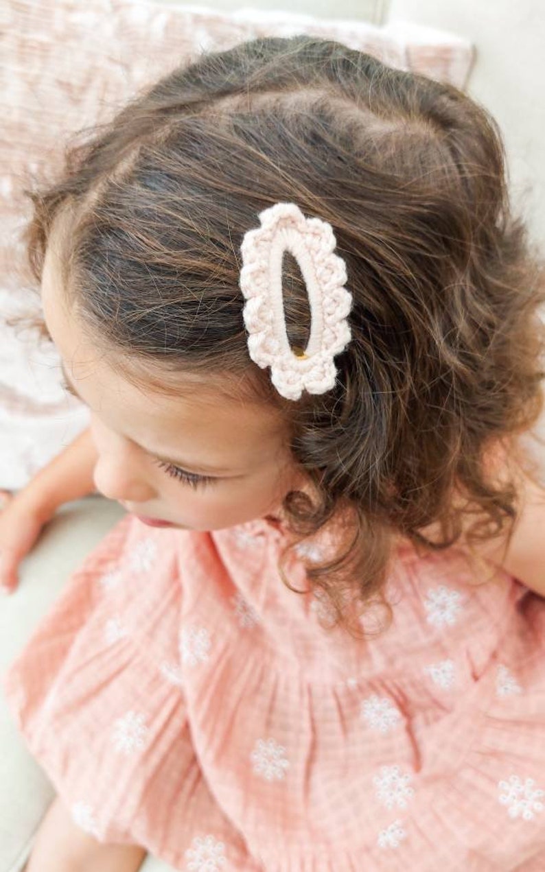 May include: A young girl with curly brown hair wearing a pink dress with white floral embroidery. She has a pink crocheted hair clip in her hair.