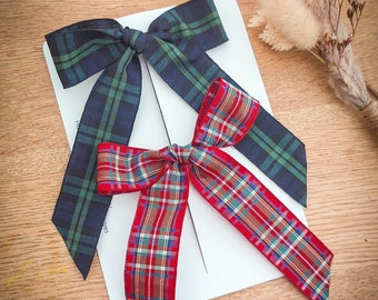 May include: Two plaid fabric bows in the image. One is red with green and white accents, and the other is navy blue with green accents. The bows are on a white card, with dried flowers in the background. The bows are on a wooden surface.