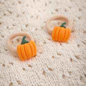 May include: Two orange pumpkin-shaped hair ties with green stems. The pumpkin charms are attached to beige elastic bands. The hair accessories are set against a cream-colored, textured fabric background.