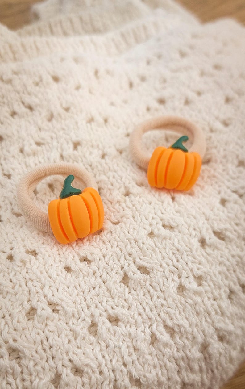 May include: Two orange pumpkin-shaped hair ties with green stems. The pumpkin charms are attached to beige elastic bands. The hair accessories are set against a cream-colored, textured fabric background.