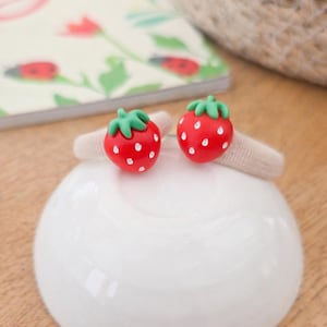 May include: Two red strawberry hair clips with white dots and green stems sit on a beige hair tie. The clips rest on a white, round, ceramic dish. A notebook with floral illustrations is in the background.