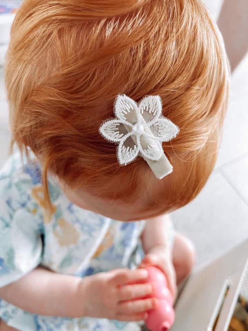 May include: A white flower hair clip with a pearl center. The clip is in a young person's red hair.