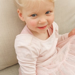 May include: A toddler with blonde hair styled in two high pigtails, each secured with a small white bow. The child is wearing a long-sleeved, light pink shirt layered under a matching pink dress featuring a multi-layered, ruffled tulle skirt.