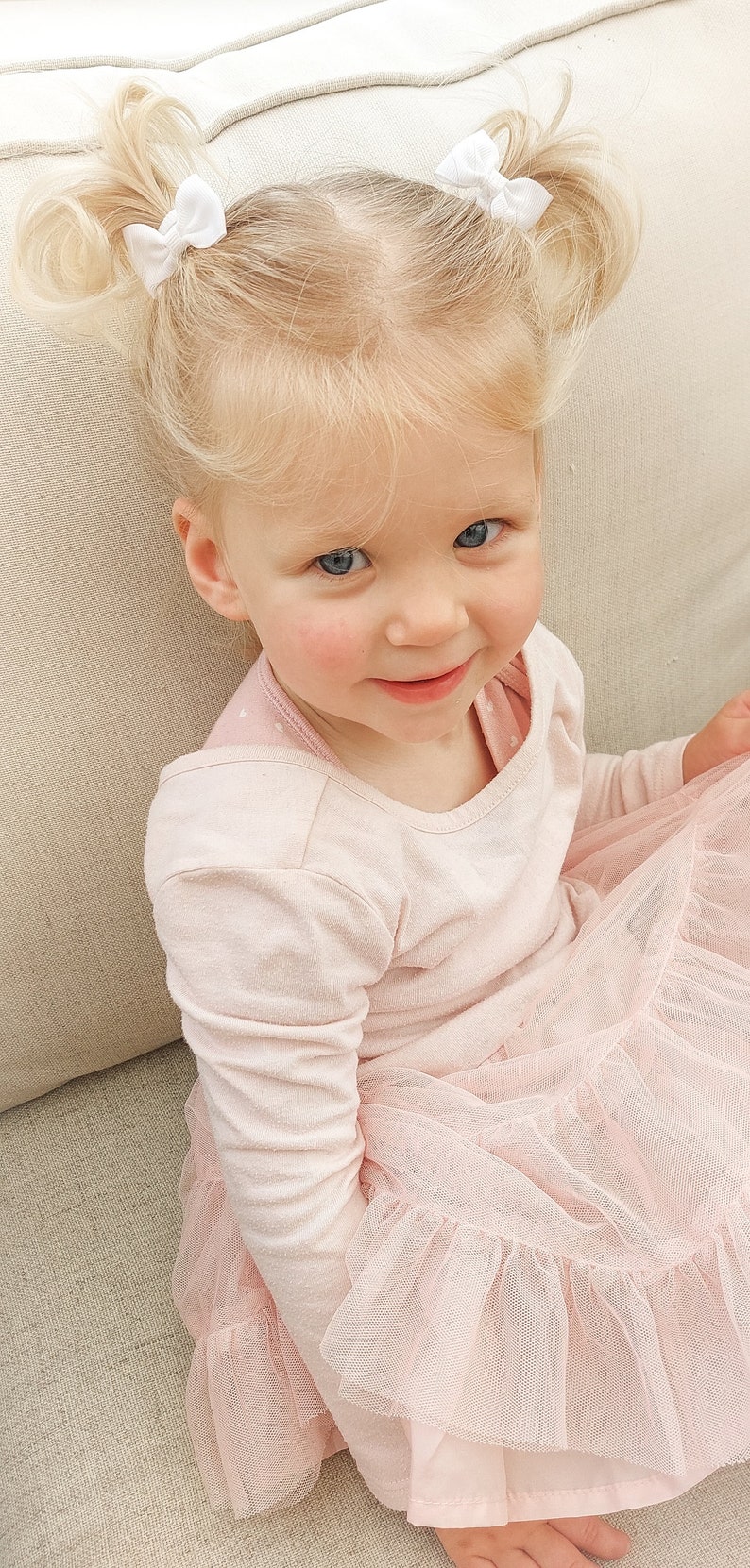 May include: A toddler with blonde hair styled in two high pigtails, each secured with a small white bow. The child is wearing a long-sleeved, light pink shirt layered under a matching pink dress featuring a multi-layered, ruffled tulle skirt.