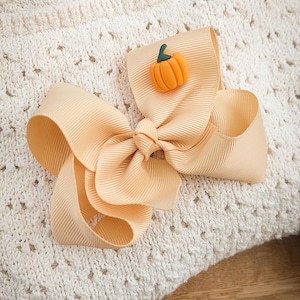 May include: A large, peach-colored hair bow with a small orange pumpkin embellishment. The bow is made of a ribbed fabric and is set against a cream-colored, textured background. The pumpkin has a green stem.