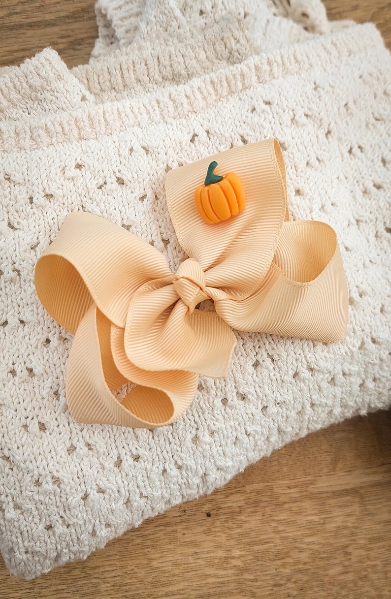 May include: A large, peach-colored hair bow with a small orange pumpkin embellishment. The bow is made of a ribbed fabric and is set against a cream-colored, textured background. The pumpkin has a green stem.