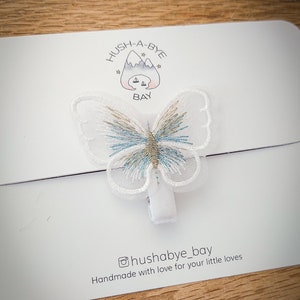K&ouml;nnte beinhalten: Wei&szlig;er Schmetterlings-Haarklipp mit blauer und goldener Stickerei. Der Clip befindet sich auf einer wei&szlig;en Karte mit dem Text "HUSH-A-BYE BAY" und "@hushabye_bay Handmade with love for your little loves".