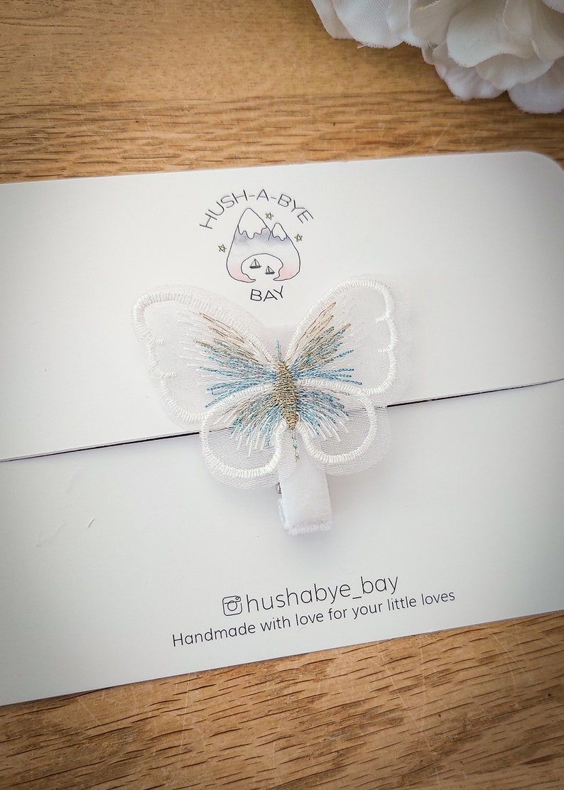 K&ouml;nnte beinhalten: Wei&szlig;er Schmetterlings-Haarklipp mit blauer und goldener Stickerei. Der Clip befindet sich auf einer wei&szlig;en Karte mit dem Text "HUSH-A-BYE BAY" und "@hushabye_bay Handmade with love for your little loves".
