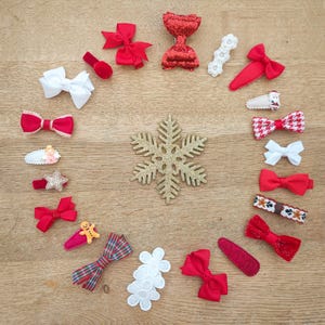 May include: A collection of 18 red and white hair bows with various designs, including plaid, glitter, and snowflakes. The bows are arranged in a circle around a gold snowflake on a wooden surface.