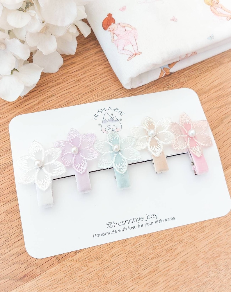May include: Set of four pastel colored flower hair clips with pearl accents. The clips are on a white card with the text "Hush-A-Bye" and "@hushabye_boy Handmade with love for your little loves".