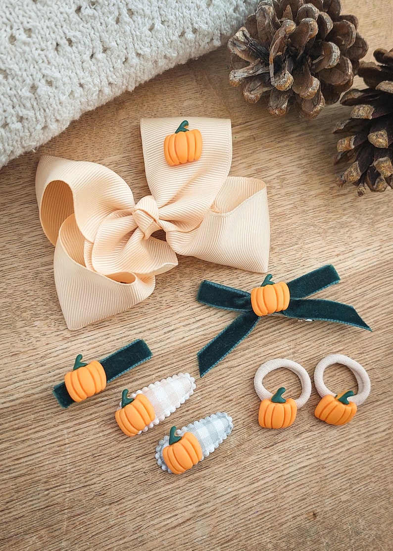 May include: A collection of fall-themed hair accessories, including a large peach-colored bow, green velvet barrettes, and hair ties. Each item is adorned with a small orange pumpkin with a green stem. The accessories are arranged on a wooden surface.