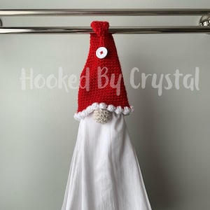 Puede incluir: Sombrero de gnomo de punto rojo con un pomp&oacute;n blanco y un bot&oacute;n rojo. El sombrero est&aacute; colgado en un toallero con una toalla de cocina blanca.