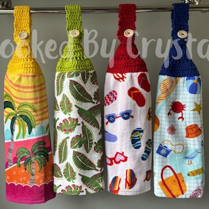 May include: Four hanging kitchen towels with crochet tops and button closures. The towels feature colorful beach-themed patterns, including palm trees, tropical leaves, and beach accessories. The crochet tops are yellow, green, red, and blue.