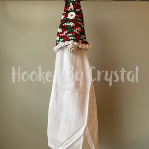 Puede incluir: Una toalla de cocina blanca con un gorro de gnomo tejido a crochet en rayas rojas, verdes y blancas. El gorro tiene un pomp&oacute;n blanco y un bot&oacute;n blanco.