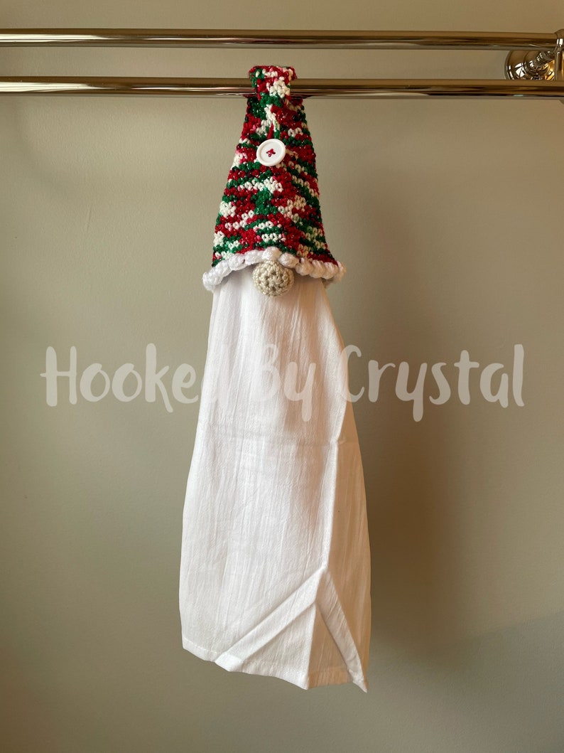 Puede incluir: Una toalla de cocina blanca con un gorro de gnomo tejido a crochet en rayas rojas, verdes y blancas. El gorro tiene un pomp&oacute;n blanco y un bot&oacute;n blanco.