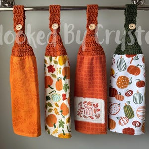 May include: Four hanging kitchen towels with crocheted tops and wooden buttons. The first is solid orange with a leaf texture. The second features a pumpkin and leaf print. The third is orange with a patch reading Happy Fall. The fourth has a green top and pumpkin print.