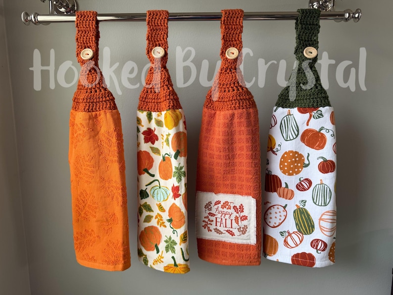May include: Four hanging kitchen towels with crocheted tops and wooden buttons. The first is solid orange with a leaf texture. The second features a pumpkin and leaf print. The third is orange with a patch reading Happy Fall. The fourth has a green top and pumpkin print.
