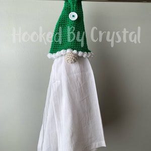 Puede incluir: Un gorro de gnomo de crochet verde con un bot&oacute;n blanco y un pomp&oacute;n blanco en la parte superior. El gorro est&aacute; colgado en un toallero con una toalla de cocina blanca colgando debajo. El texto "Hooked By Crystal" es visible en el fondo.