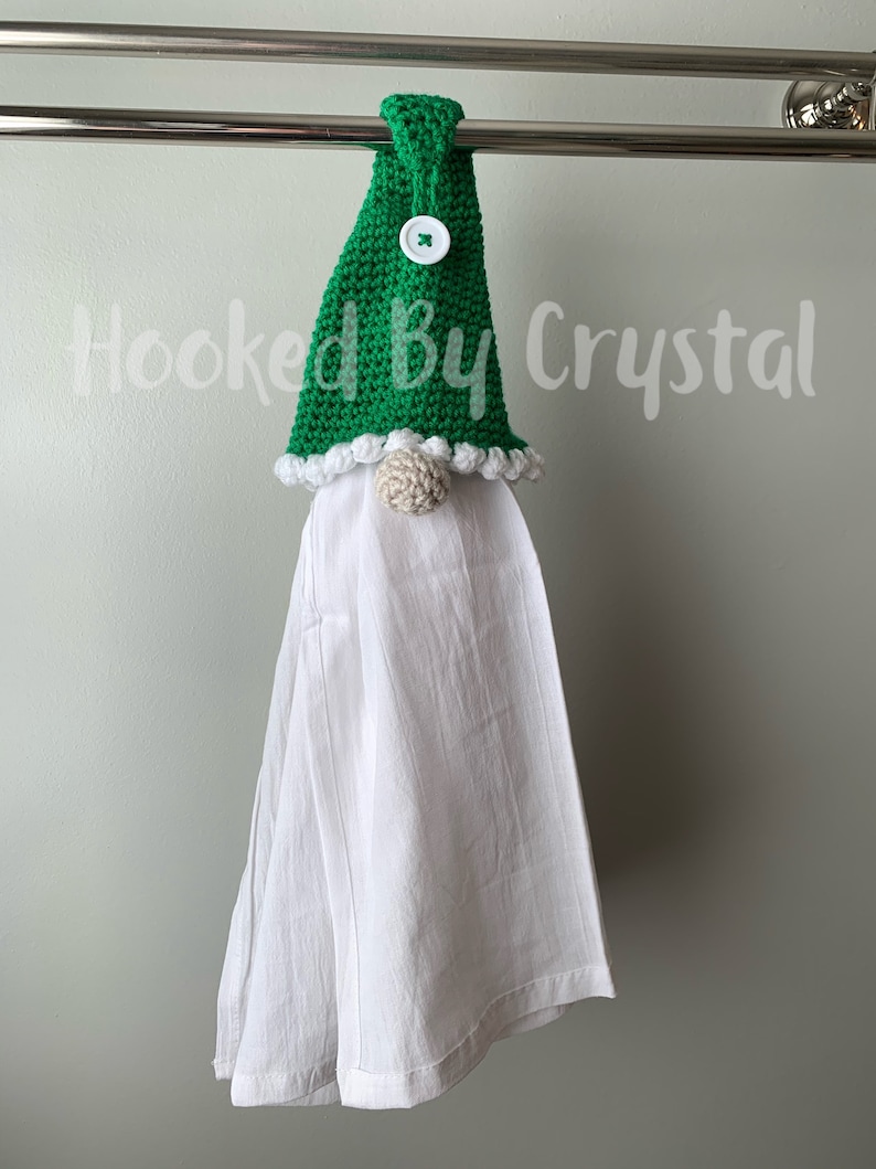 Puede incluir: Un gorro de gnomo de crochet verde con un bot&oacute;n blanco y un pomp&oacute;n blanco en la parte superior. El gorro est&aacute; colgado en un toallero con una toalla de cocina blanca colgando debajo. El texto "Hooked By Crystal" es visible en el fondo.