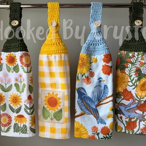 May include: Four hanging kitchen towels with crocheted tops in dark green, yellow, and light blue, each secured with a wooden button. The towels feature floral patterns, a yellow gingham print with a sunflower square, and blue birds on branches.