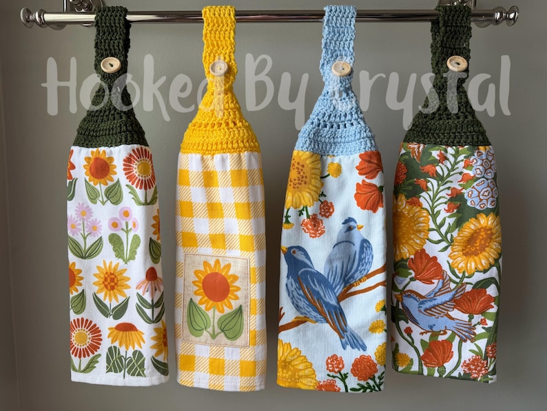 May include: Four hanging kitchen towels with crocheted tops in dark green, yellow, and light blue, each secured with a wooden button. The towels feature floral patterns, a yellow gingham print with a sunflower square, and blue birds on branches.