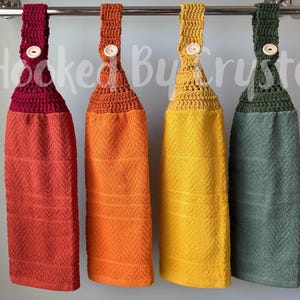 May include: Four hanging kitchen towels with crocheted tops in burgundy, orange, yellow, and olive green. Each towel features a matching crocheted loop secured with a light-colored button, hanging from a silver rod. The text Hooked By Crystal is visible.
