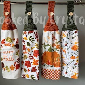 May include: Four hanging kitchen towels with crocheted tops in orange and dark green, each secured with a light-colored button. The towels feature autumn-themed prints including fall leaves, pumpkins, and the text Happy Fall on a white background.
