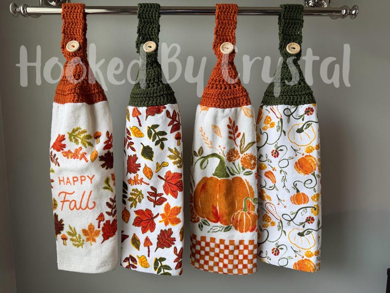 May include: Four hanging kitchen towels with crocheted tops in orange and dark green, each secured with a light-colored button. The towels feature autumn-themed prints including fall leaves, pumpkins, and the text Happy Fall on a white background.