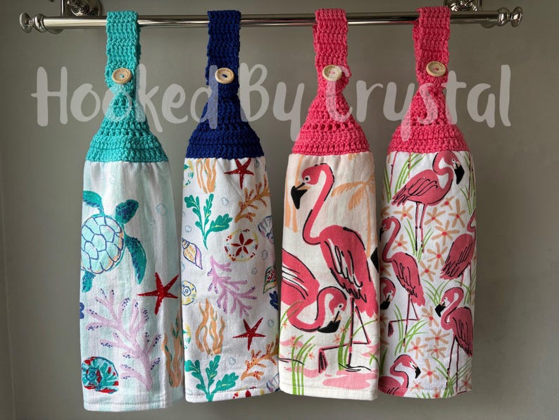 May include: Four hanging kitchen towels with crochet tops and wooden buttons. The towels feature sea turtle, starfish, coral, and flamingo designs. The text "Hooked By Crystal" is visible in the background.