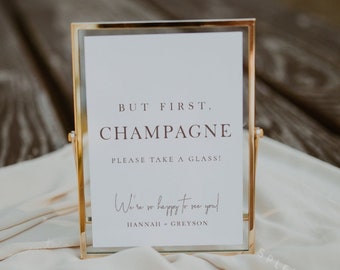 May include: A gold-framed sign with the text "BUT FIRST. CHAMPAGNE PLEASE TAKE A GLASS!" and "We're so happy to see you! HANNAH & GREYSON" in a cursive font.