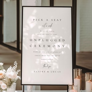 May include: A framed sign with the text "Pick a seat, not a side. Unplugged Ceremony." is displayed at an outdoor event. The sign is on a white table with floral arrangements and candles. The sign is black and white.