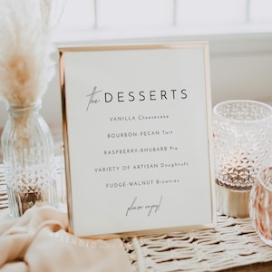 May include: A gold framed sign with the text "the DESSERTS" and a list of desserts: Vanilla Cheesecake, Bourbon-Pecan Tart, Raspberry-Rhubarb Pie, Variety of Artisan Doughnuts, Fudge-Walnut Brownies. The sign is on a table with a woven placemat and two glass candle holders.