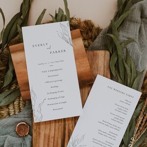 Könnte beinhalten: Zwei weiße Hochzeitskarten mit schwarzem Text und botanischen Illustrationen liegen auf einem Holzbrett, umgeben von Grün und einem grauen Tuch. Die Karten listen die Zeremoniedetails und die Namen der Hochzeitsgesellschaft auf.