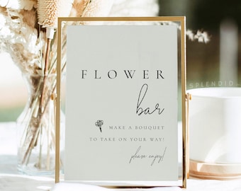 May include: A gold-framed sign with the text "FLOWER bar" and the words "Make a bouquet to take on your way! Please enjoy!"  The sign is sitting on a table with a vase of dried flowers in the background.