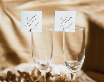 May include: Two champagne flutes with place cards for a wedding reception. The place cards are white with black text and say "Mr. James Thompson" and "Mrs. Alexandra Walker".