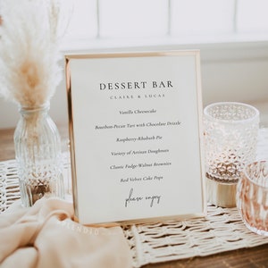 May include: A dessert menu in a gold-framed display, listing desserts such as vanilla cheesecake and raspberry-rhubarb pie. The menu is placed on a table with decorative items, including a glass vase with pampas grass and two candle holders. The text reads "DESSERT BAR".