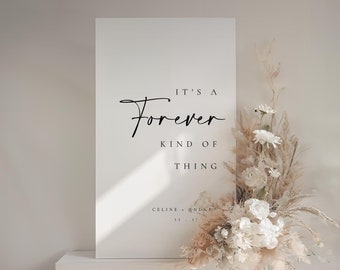 May include: White sign with black text that reads "It's a Forever Kind of Thing" with the names "Celine & Andrew" below. The sign is leaning against a white pedestal and a bouquet of dried flowers is in front of it.