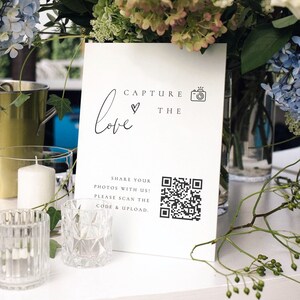 May include: White sign with black text that reads "Capture the love" with a heart and camera icon. A QR code is below the text with instructions to share photos with the code. The sign is on a table with flowers and candles.