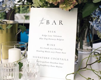 May include: A white sign with black text that reads "the BAR" and lists beer, wine, and signature cocktails. The sign is sitting on a white table with candles and flowers.