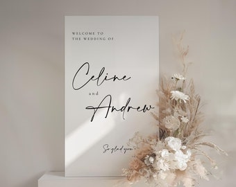 May include: A minimalist wedding sign in white, featuring the names "Celine and Andrew" in cursive script. The sign is decorated with a floral arrangement of white and beige flowers and pampas grass. The text "Welcome to the wedding of" is above the names.