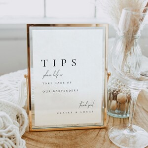 Könnte beinhalten: Ein goldgerahmtes Schild mit dem Wort „TIPS“ in einer minimalistischen Schriftart. Auf dem Schild steht „please help us take care of our bartenders“ und „thank you!“ mit Namen darunter. Ein Weinglas und eine dekorative Vase sind im Hintergrund.