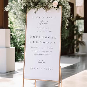 May include: A wedding ceremony sign with a white background and gold metal frame. The sign is decorated with greenery and a single pink rose. Text on the sign reads "Pick a seat not a side" and "Unplugged Ceremony".