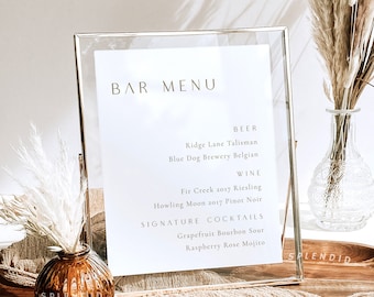 May include: A gold framed bar menu sign with a white background and gold lettering. The menu lists beer, wine, and signature cocktails. The sign is sitting on a wooden table with a brown fabric runner.